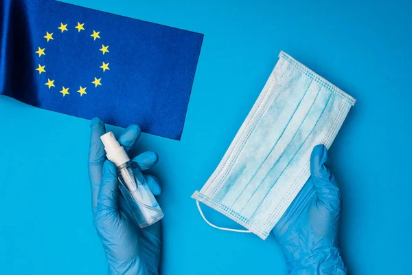 Vista superior del médico sosteniendo máscara médica y botella de desinfectante de manos cerca de la bandera de la unidad europea sobre fondo azul - foto de stock