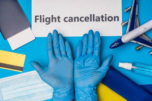 Vista superior del médico en guantes de látex cerca de la tarjeta con cancelación de vuelo, avión de juguete con bandera de Ucrania y máscara médica sobre fondo azul - foto de stock