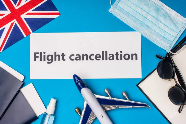 Top view of card with flight cancellation lettering near flag of United Kingdom, toy airplane and medical mask on blue surface — стоковое фото