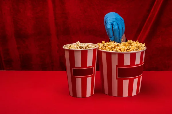 Ausgeschnittene Ansicht einer Frau im Latex-Handschuh, die Popcorn aus einem Eimer auf roter Fläche mit Velours im Hintergrund nimmt — Stockfoto