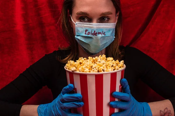 Frau in medizinischer Maske mit Epidemie-Schriftzug und Latexhandschuhen hält Eimer mit Popcorn mit rotem Velours im Hintergrund — Stockfoto