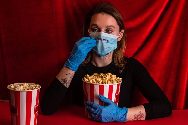 Frau in Latexhandschuhen und medizinischer Maske mit Epidemie-Schriftzug hält Popcorn auf roter Oberfläche mit Velours im Hintergrund — Stockfoto