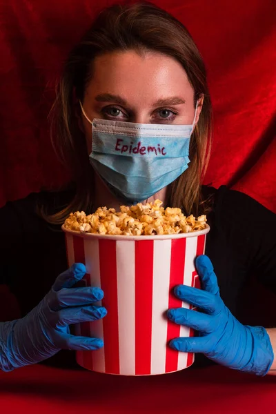 Junge Frau in medizinischer Maske mit Epidemie-Schriftzug hält Eimer mit Popcorn auf roter Fläche mit Velours im Hintergrund — Stockfoto