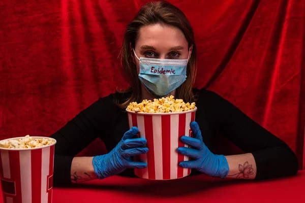 Frau in Latexhandschuhen und medizinischer Maske mit Epidemie-Schriftzug hält Eimer mit Popcorn auf roter Oberfläche mit Velours im Hintergrund — Stockfoto