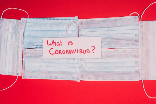 Visão superior do cartão com o que é coronavírus lettering em máscaras médicas no fundo vermelho — Fotografia de Stock