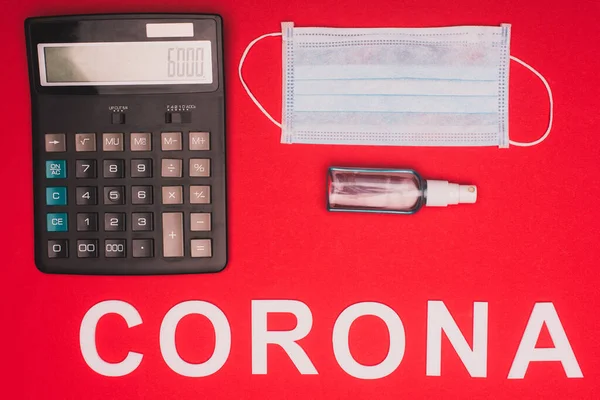 Top view of calculator with medical mask and bottle of hand sanitizer near corona lettering on red background — Stock Photo