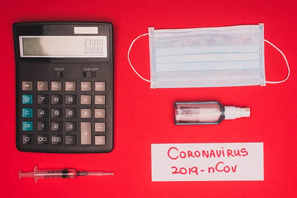 Top view of medical mask, calculator near syringe and card with coronavirus 2019-nCov lettering on red background — Stock Photo