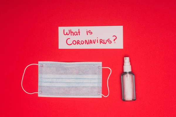 Visão superior do cartão com o que é coronavírus lettering perto de higienizador de mão e máscara médica na superfície vermelha — Fotografia de Stock