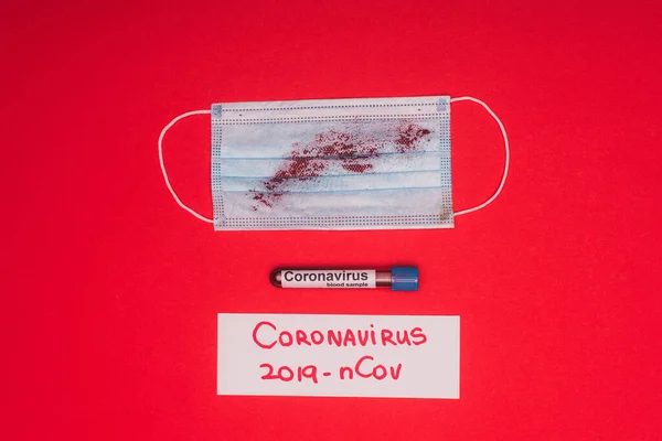 Top view of medical mask with blood near test tube with blood sample and card with coronavirus 2019-nCov lettering on red background — Stock Photo