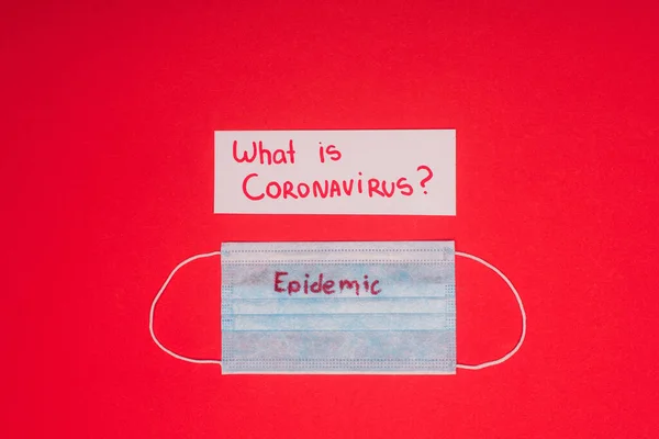 Vista superior do cartão com o que é coronavírus e máscara médica com lettering epidêmico isolado no vermelho — Fotografia de Stock