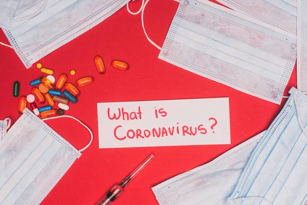 Top view of card with what is coronavirus lettering near medical masks, pills and syringe on red background — Stock Photo