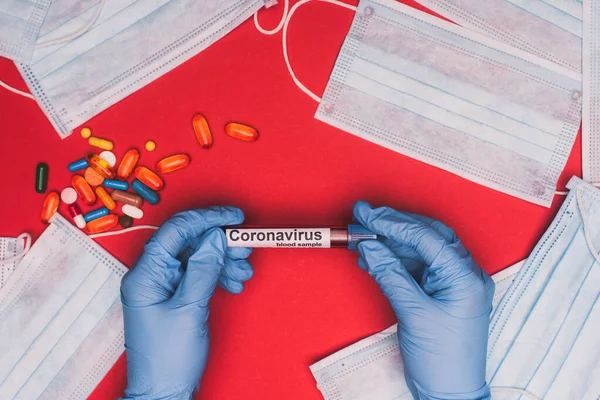 Vue du dessus du médecin tenant un tube à essai avec échantillon de sang et lettrage coronavirus près des pilules et des masques médicaux sur fond rouge — Photo de stock