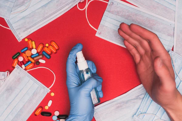 Vue du dessus de l'homme en gant de latex à l'aide d'un désinfectant pour les mains près des masques médicaux avec des pilules sur fond rouge — Photo de stock