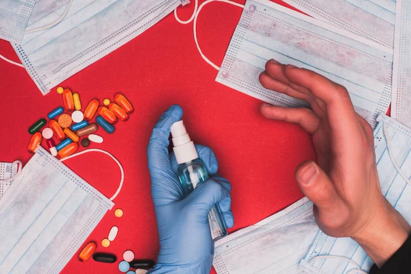 Vue recadrée de l'homme tenant une bouteille de désinfectant pour les mains près des pilules et des masques médicaux sur la surface rouge — Photo de stock