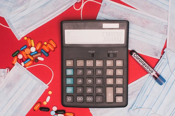 Top view of calculator, test tube with blood sample and coronavirus lettering near medical masks and pills on red surface — Stock Photo