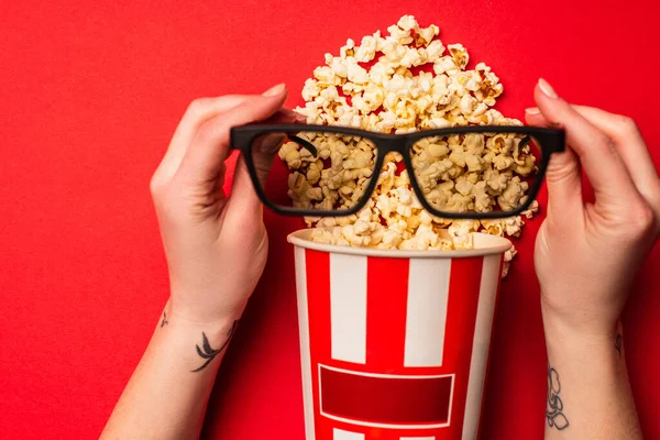 Top view of girl holding sunglasses near cardboard bucket with popcorn on red background — Stock Photo