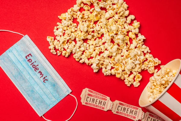 Top view of medical mask with epidemic lettering, popcorn and cinema tickets on red background — Stock Photo