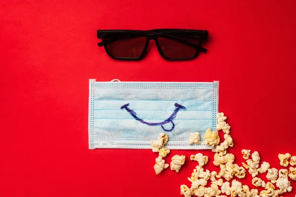 Vista superior de gafas de sol cerca de máscara médica con sonrisa y palomitas de maíz en la superficie roja - foto de stock