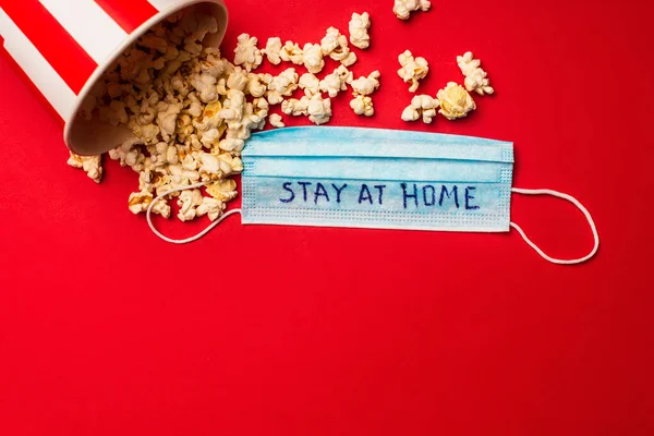 Vista superior de cubo con palomitas de maíz y máscara médica con letras de estancia en casa sobre fondo rojo - foto de stock