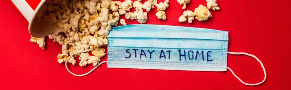 Top view of medical mask with stay at home lettering and popcorn on red surface, panoramic shot — Stock Photo