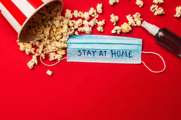 Top view of medical mask with stay at home lettering near hand sanitizer and popcorn on red background — Stock Photo