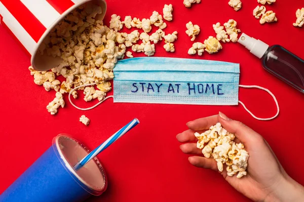 Top view of woman holding popcorn near medical mask with stay at home lettering, hand disanitizer and paper cup on red background — стоковое фото