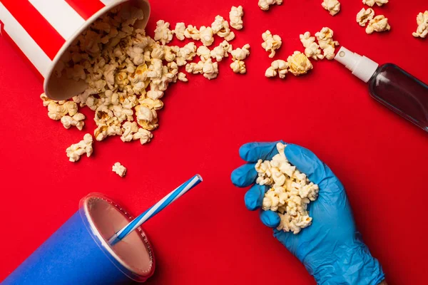 Vue du dessus de la personne en gant de latex tenant du pop-corn près d'un désinfectant pour les mains et d'une tasse en papier sur fond rouge — Stock Photo