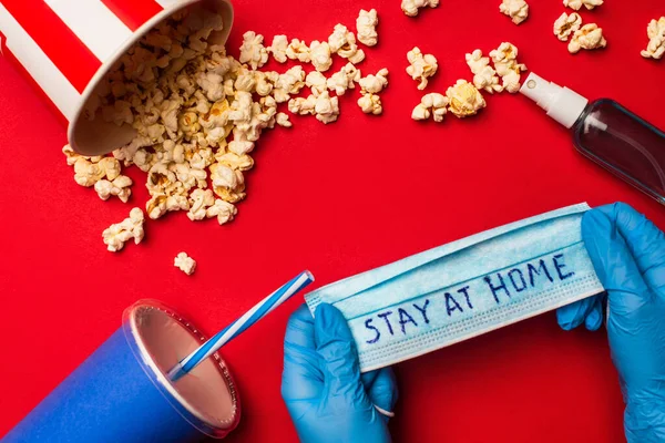 Top view of person holding medical mask with stay at home lettering near hand sanitizer and popcorn on red surface — Stock Photo
