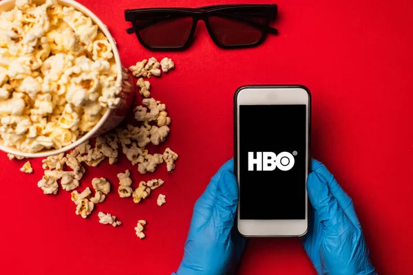 KYIV, UKRAINE - MARCH 26, 2020: Top view of person holding smartphone with hbo app near sunglasses and popcorn on red background — Stock Photo