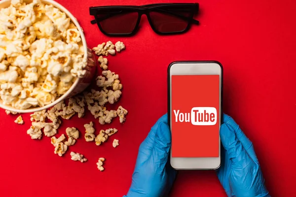 KYIV, UKRAINE - MARCH 26, 2020: Top view of person holding smartphone with youtube app near popcorn and sunglasses on red background — Stock Photo
