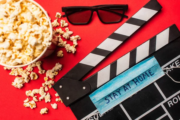 Top view of clapperboard with medical mask and stay at home lettering near popcorn and sunglasses on red surface — Stock Photo