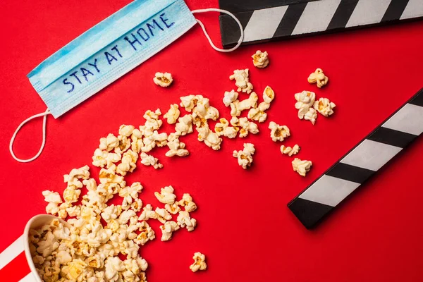 Vue du dessus du pop-corn près de clapperboard et masque médical avec lettrage séjour à la maison sur fond rouge — Photo de stock