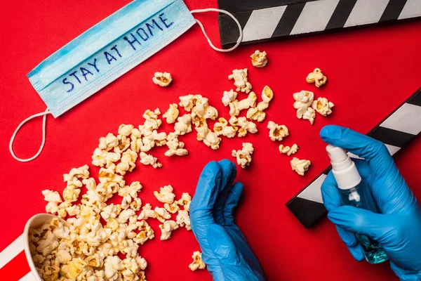 Vista superior de la persona que usa desinfectante de manos cerca de palomitas de maíz y máscara médica con letras de estancia en casa sobre fondo rojo - foto de stock