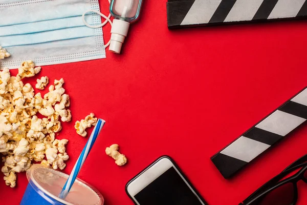 Top view of popcorn, smartphone with medical mask and hand disanitizer on red surface — стоковое фото