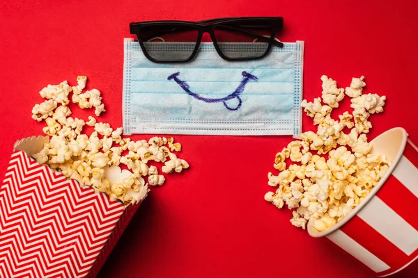 Draufsicht auf medizinische Maske mit Lächeln in der Nähe von Sonnenbrille und Eimern mit Popcorn auf roter Oberfläche — Stockfoto