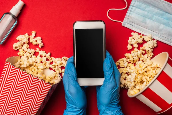 Vista superior de la persona que sostiene el teléfono inteligente con pantalla en blanco cerca de la máscara médica y palomitas de maíz en la superficie roja - foto de stock