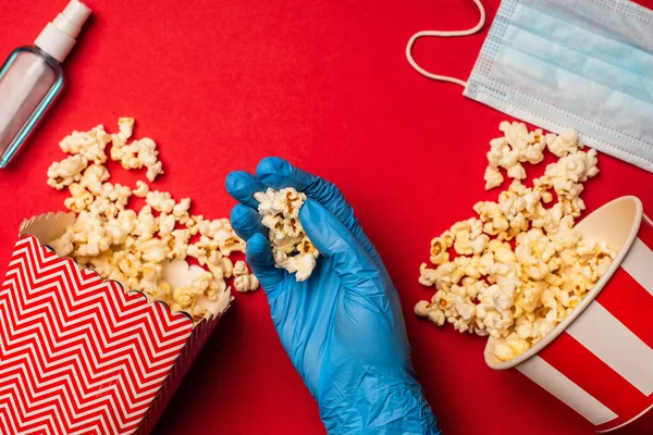 Draufsicht der Person in Latex-Handschuh mit Popcorn in der Nähe Händedesinfektionsmittel und medizinische Maske auf rotem Hintergrund — Stockfoto