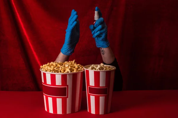 Ausgeschnittene Ansicht einer Frau in Latex-Handschuhen mit Händedesinfektionsmittel in der Nähe von Eimern mit Popcorn auf roter Oberfläche mit Velours im Hintergrund — Stockfoto