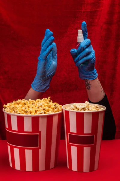 Обрезанный вид попкорна рядом с девушкой в латексных перчатках с помощью дезинфицирующего средства для рук на красной поверхности с велюром на заднем плане — стоковое фото