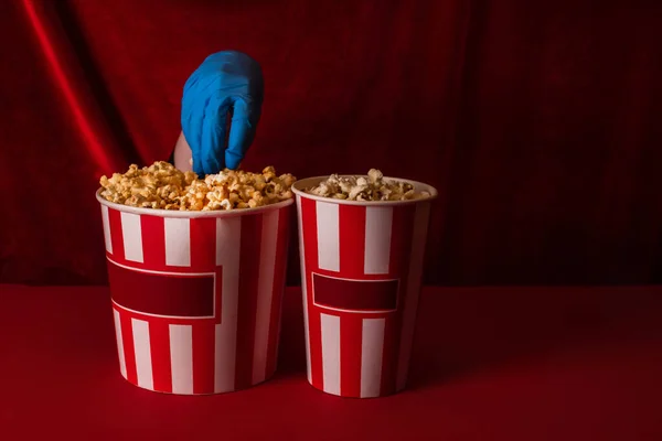 Cropped view of woman in latex glove taking popcorn from bucket on red surface and velour at background — Stock Photo