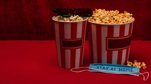 Seaux avec pop-corn près des lunettes de soleil et masque médical avec lettrage de séjour à la maison sur la surface rouge et velours à l'arrière-plan — Photo de stock