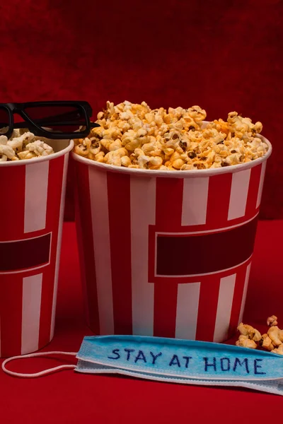 Medical mask with stay at home lettering near buckets with popcorn and sunglasses on red surface and velour at background — Stock Photo