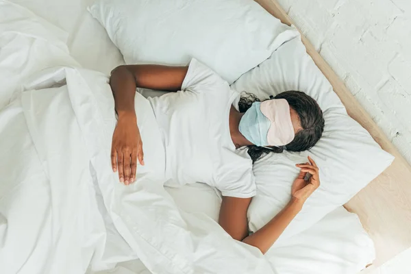 Vista dall'alto della ragazza afroamericana in maschera del sonno e maschera medica dormire a letto — Foto stock