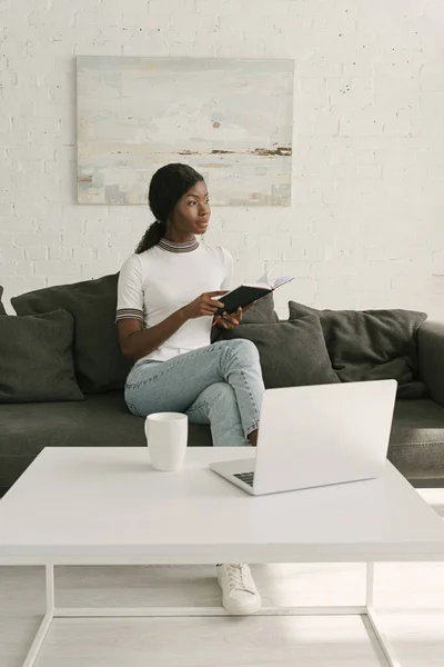 Reflexivo freelancer afroamericano sosteniendo portátil mientras está sentado en el sofá cerca de la mesa con el ordenador portátil - foto de stock