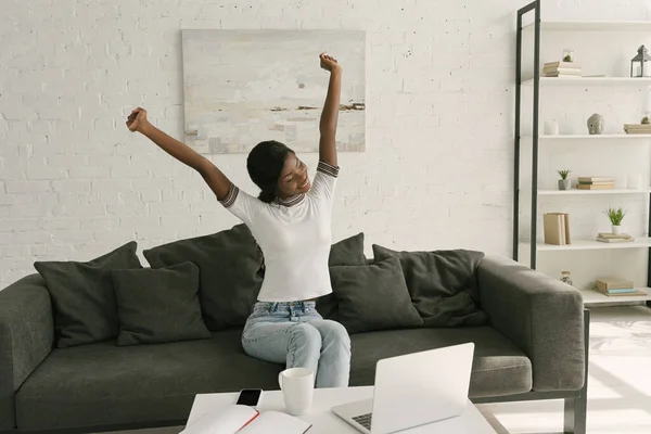 Souriant afro-américain pigiste étirement avec les mains levées tout en étant assis sur le canapé près de la table avec ordinateur portable — Photo de stock