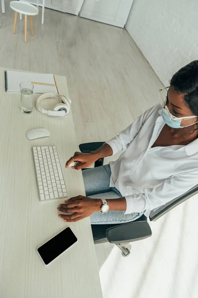 Overhead view of african american freelancer in medical mask spraying antiseptic on keyboard near smartphone and wireless headphones — Stock Photo