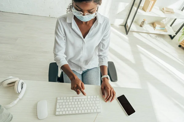 Visão aérea do freelancer afro-americano em máscara médica pulverizando anti-séptico no teclado — Fotografia de Stock