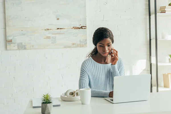 Freelancer afro-americano atencioso falando no smartphone perto de laptop, fones de ouvido sem fio e xícara de café — Fotografia de Stock