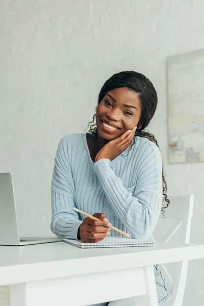 Felice freelance africano americano sorridente alla macchina fotografica mentre tiene la matita vicino al notebook e laptop — Foto stock