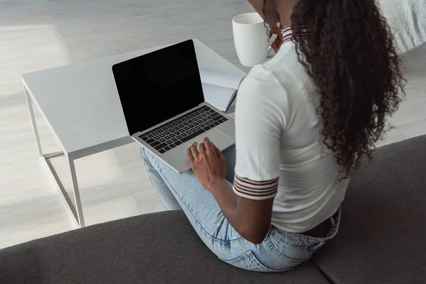 Vista ad alto angolo della ragazza afro-americana utilizzando il computer portatile con schermo bianco e tenendo in mano la tazza di caffè — Foto stock